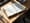 An open ledger book on a sunlit desk with handwritten numbers, and a hand of playing cards tucked underneath.